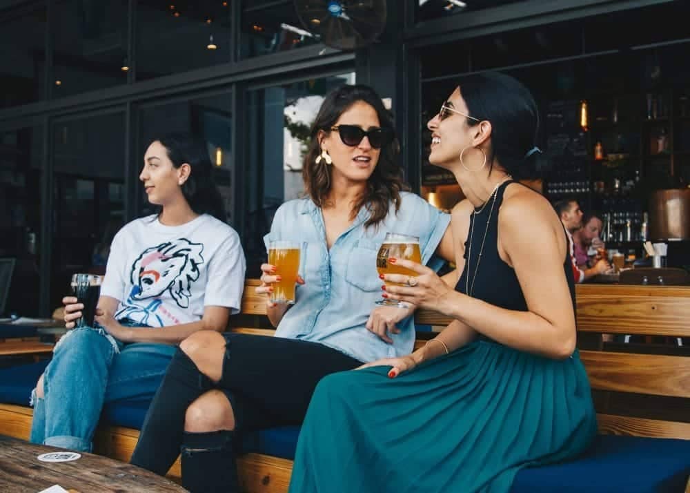 mom sits on restaurant bench chatting and drinking beers while daughter looks away